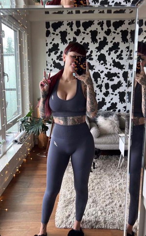 Red-haired woman with tattoos taking mirror selfie in grey activewear.