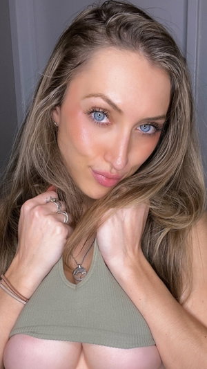 Woman with long blonde hair and blue eyes wearing olive green crop top, posing indoors.