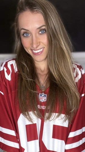 Blonde woman smiling in red 49ers jersey indoors.