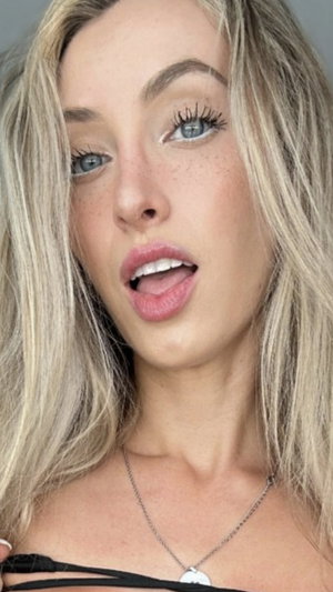 Blonde woman with blue eyes and freckles, wearing a black top and necklace, close-up selfie.