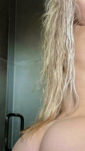 Blonde woman with wet hair, bare shoulder, standing near glass shower door.
