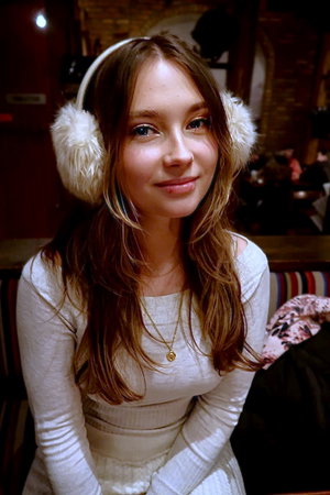 Woman with long brown hair wearing white earmuffs and sweater indoors.