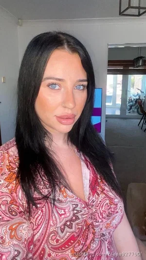 Woman with long black hair and blue eyes wearing a pink patterned top indoors.