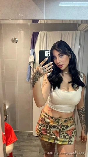 Woman with tattoos in white crop top and patterned shorts, taking mirror selfie in bathroom.