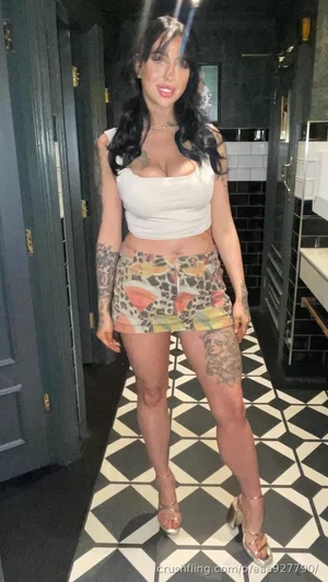 Woman with tattoos in white crop top and patterned shorts, standing in a tiled hallway.
