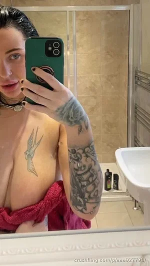Woman with tattoos taking mirror selfie in bathroom, partially covered by red towel.