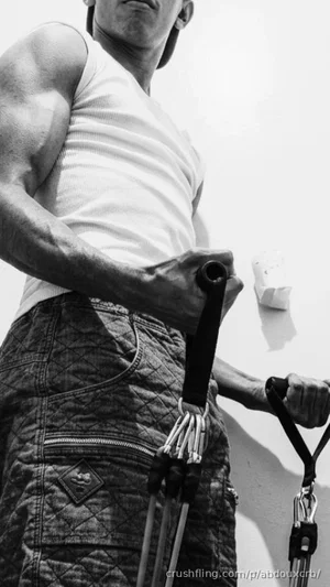 Muscular man in white tank top and cargo shorts holding climbing gear outdoors.