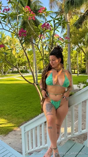Woman in green bikini with tattoos, standing on porch steps in tropical garden.