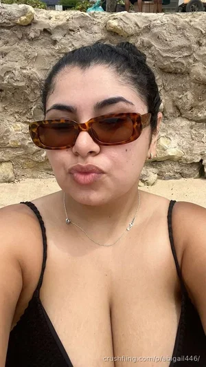 Woman with dark hair and sunglasses in a black top, outdoors near a stone wall.