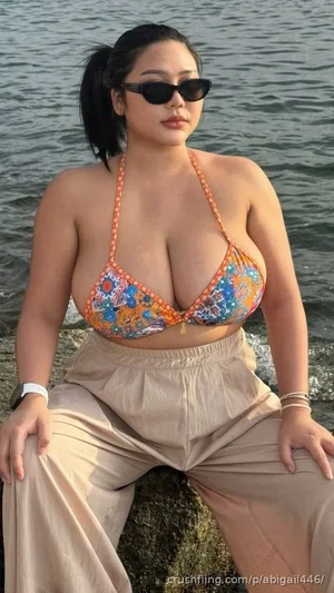 Woman in colorful bikini top and beige pants sitting by water, wearing sunglasses.