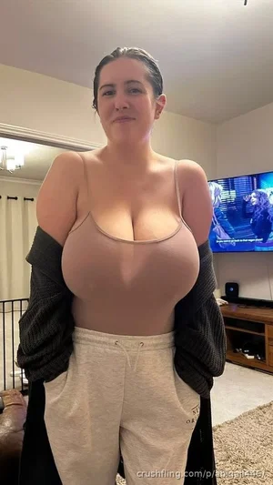 Woman in beige bra and sweatpants, standing in living room with TV on.