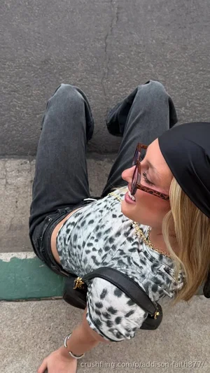 Woman with blonde hair in leopard print top and dark jeans, sitting on pavement outdoors.