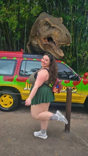 Woman in green skirt and tank top posing near Jurassic Park vehicle with dinosaur statue.
