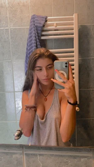 Woman with long hair in gray tank top taking mirror selfie in tiled bathroom.