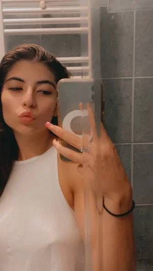 Woman in white top taking mirror selfie in bathroom, pouting lips, dark hair.