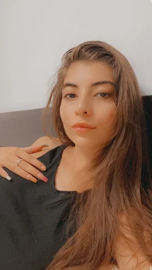 Woman with long brown hair wearing black top, sitting indoors.
