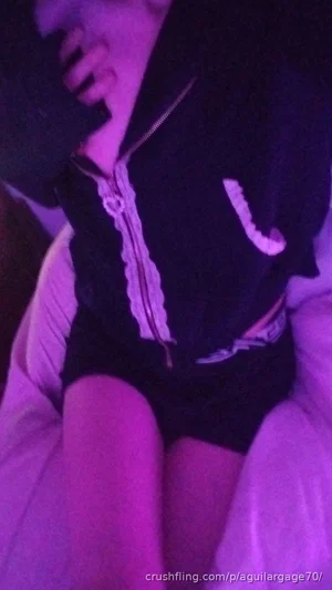Person in black zip-up top and shorts sitting on bed under purple lighting.