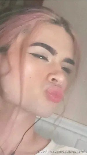 Woman with pink hair and makeup, wearing white top, making playful face indoors.