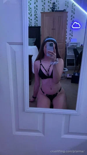Woman in black lingerie kneeling in mirror selfie, bedroom setting with green vines and neon light.