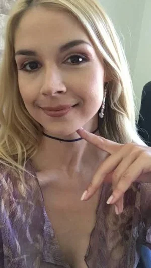 Blonde woman in purple lace top with choker, pointing at neck, indoor setting.
