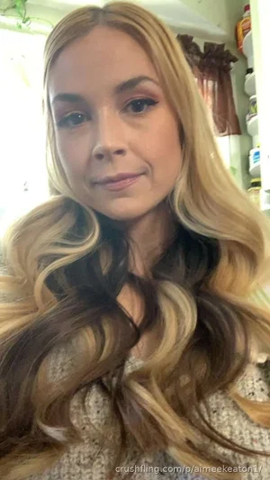 Woman with long wavy blonde hair wearing a textured top, indoors.
