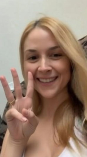 Blonde woman smiling, making peace sign, wearing white top, indoor setting.