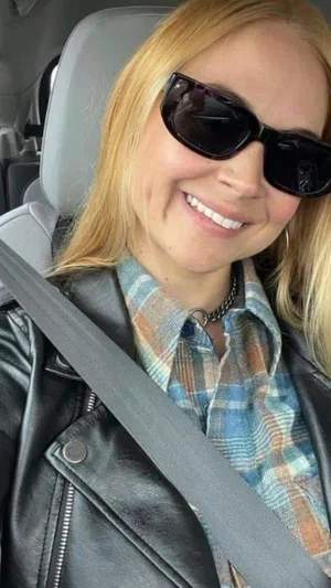 Blonde woman in plaid shirt and leather jacket, smiling in car with seatbelt.