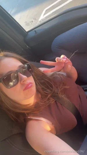 Woman with sunglasses lying in car, wearing brown top, making hand gesture.