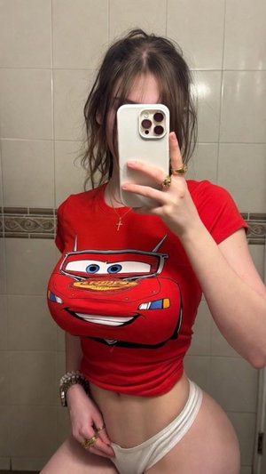 Woman in red Lightning McQueen shirt and white underwear taking mirror selfie in tiled bathroom.