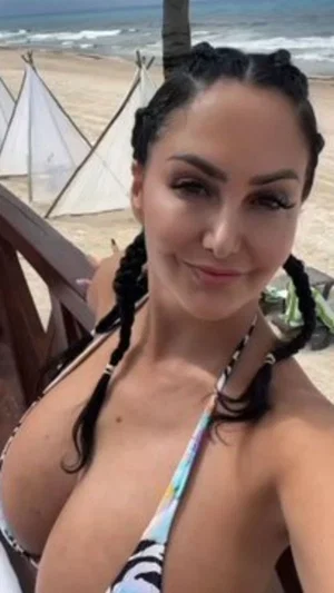 Woman in colorful bikini top at beach, braided hair, smiling selfie.