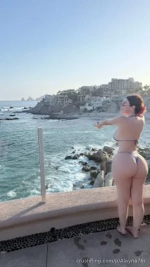 Woman in bikini standing by ocean, back view, rocky coastline in background.