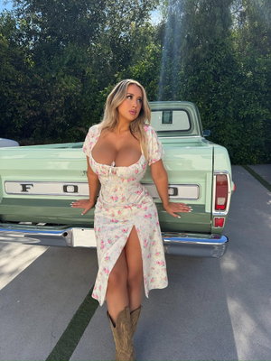 Woman in floral dress and cowboy boots posing by vintage truck outdoors.