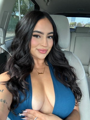Woman with long dark hair in blue top sitting in car, hand on chest.