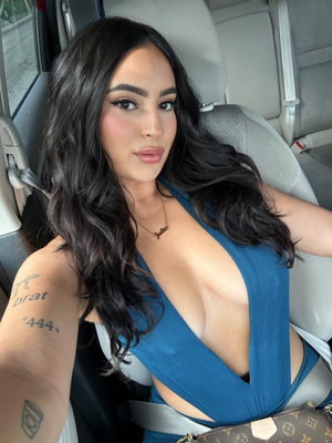 Woman with long dark hair in blue top sitting in car, showing tattoos on arm.