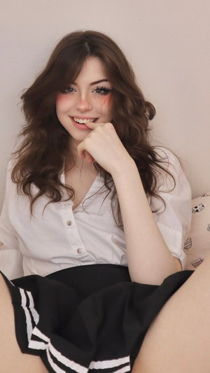 Brunette woman in white blouse and black pleated skirt sitting indoors.