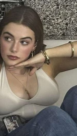 Woman with brown hair in white top and jeans, sitting indoors with hand on chin.