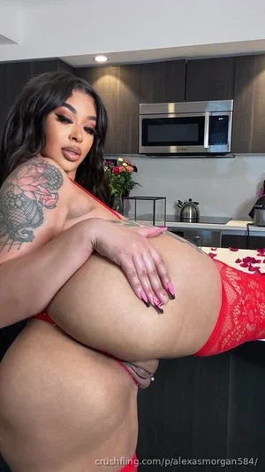 Woman with tattooed arm in red lingerie posing in kitchen.