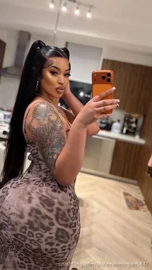 Woman with long black hair and tattoos wears leopard print dress, taking mirror selfie in kitchen.