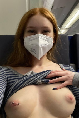 Woman with red hair and mask, top partially pulled down, showing bare breasts with piercings,