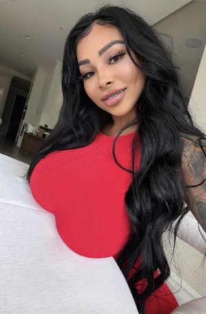 Woman with long black hair in red top, sitting on bed indoors.