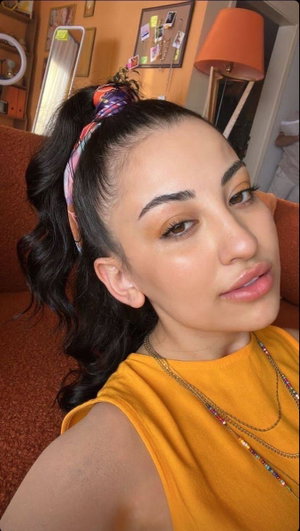 Woman with dark hair in high ponytail, wearing yellow top and layered necklaces, indoors.
