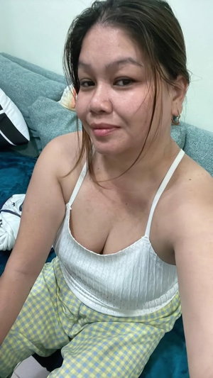 Woman in white tank top and checkered pants sitting on bed, indoor setting.