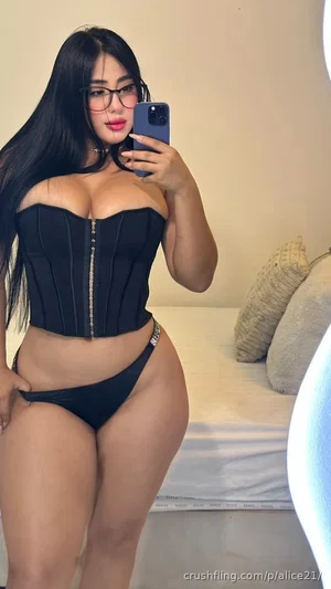 Woman with long black hair in black corset and underwear, taking mirror selfie in bedroom.