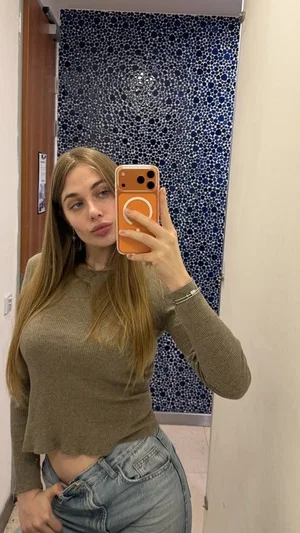 Woman with long hair in a brown crop top and jeans taking a mirror selfie in a bathroom.