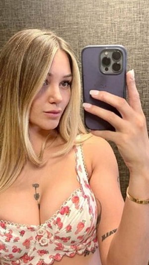 Blonde woman in pink lace bra holds phone selfie indoors.
