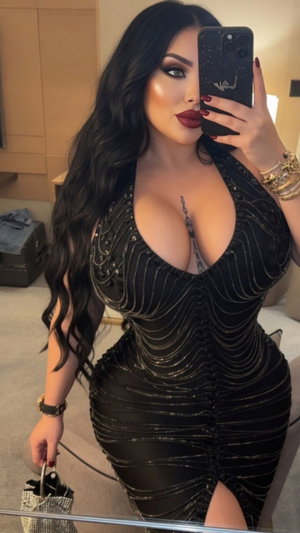 Woman with long dark hair in black sequined dress taking mirror selfie indoors.