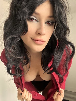 Woman with dark wavy hair, wearing red shirt open over black top, posing indoors.