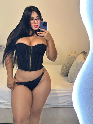 Woman with long black hair in black corset and thong, taking mirror selfie in bedroom.