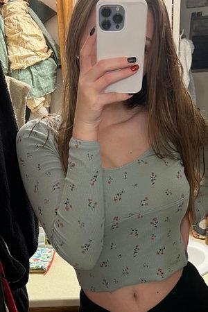 Woman in floral long-sleeve crop top and black bottoms, taking mirror selfie in bathroom.