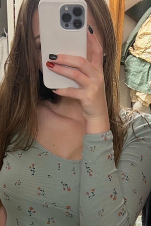 Woman with long brown hair in floral top taking mirror selfie indoors.